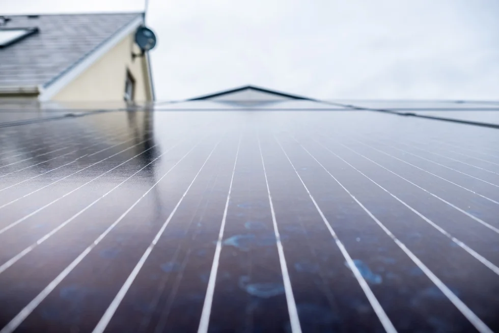 Close-up view of solar panels installed on a rooftop with a house in the background, capturing the reflective surface of the panels on an overcast day, symbolizing renewable energy and sustainable living.