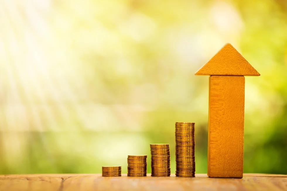 Stacks of gold coins gradually increasing in height, with a tall wooden block shaped like an arrow pointing upward, symbolizing financial growth and success, set against a warm, blurred natural background with sunlight streaming through